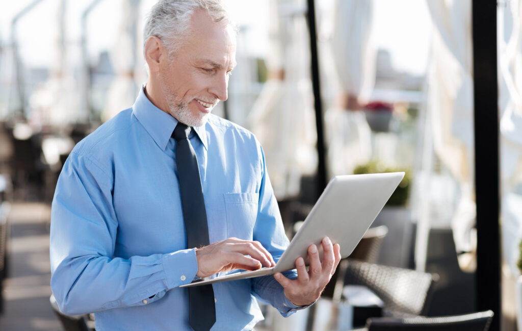 A senior professional smiling while using his laptop outdoors in a sophisticated setting. This image conveys the versatility and dependability of "fiber for business," even in dynamic environments.