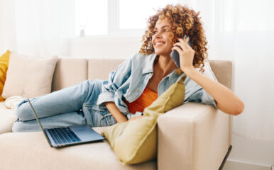 A relaxed woman lounging on a beige sofa, holding her phone while her laptop rests nearby, representing seamless "home phone connect" integration for work and communication.