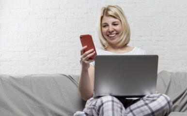 Smiling woman using her smartphone and laptop on a couch, symbolizing seamless connectivity with "business internet and phone" solutions.