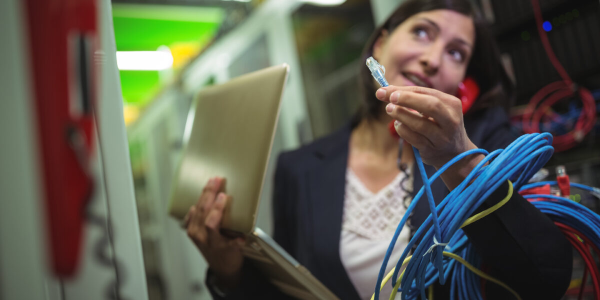 A female technician holding an Ethernet cable while multitasking on a phone and managing a laptop in a server room. The scene emphasizes the complex tasks involved in managing commercial fiber internet systems.