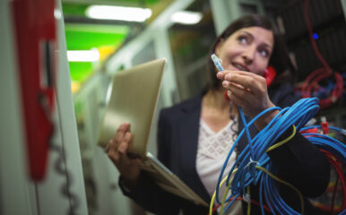 A female technician holding an Ethernet cable while multitasking on a phone and managing a laptop in a server room. The scene emphasizes the complex tasks involved in managing commercial fiber internet systems.