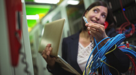 A female technician holding an Ethernet cable while multitasking on a phone and managing a laptop in a server room. The scene emphasizes the complex tasks involved in managing commercial fiber internet systems.