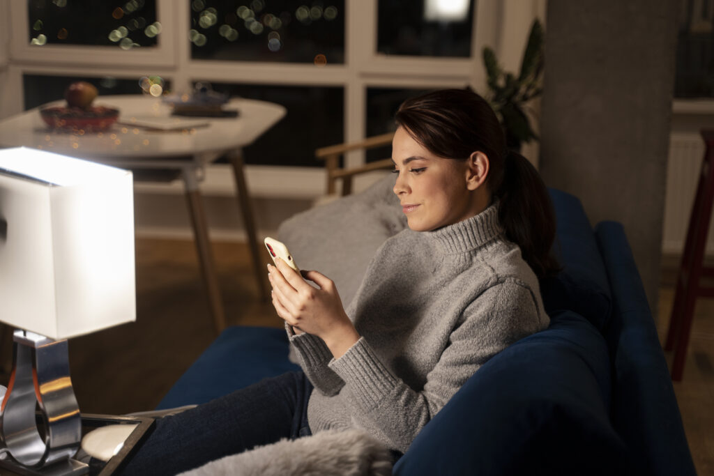 A woman comfortably using her smartphone on a navy couch at night, highlighting the importance of strong internet signal coverage in living spaces. Keywords: "best place to put wifi router in a 2 story house."