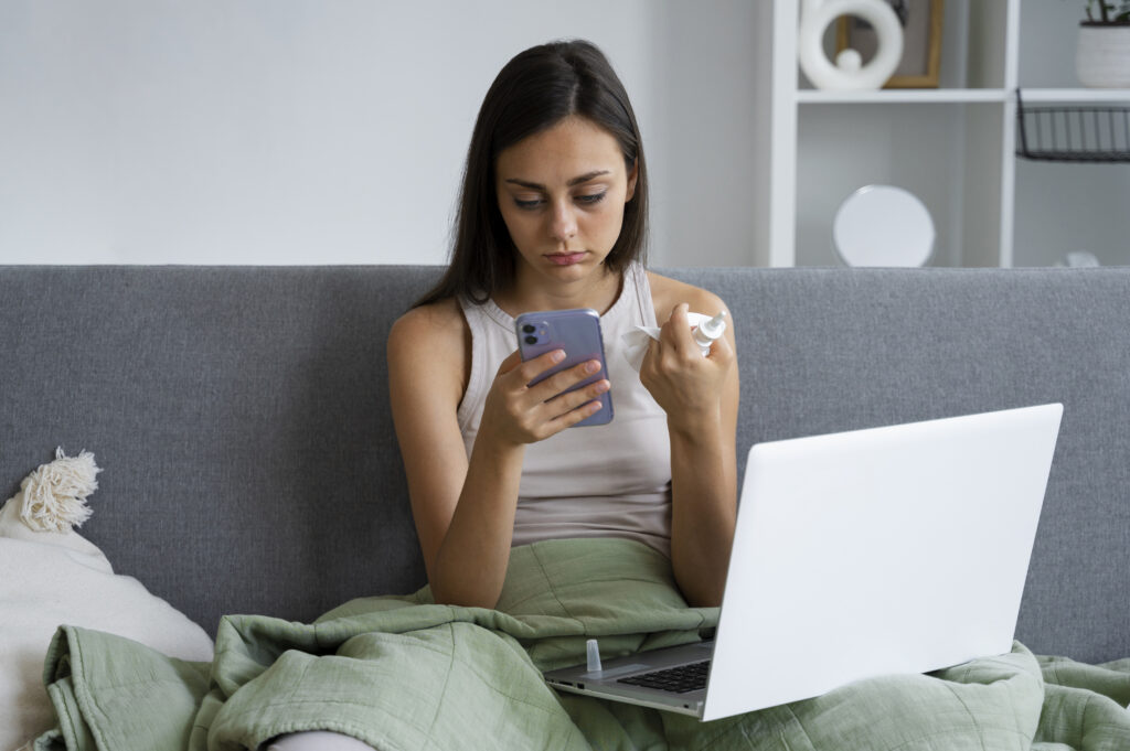 A sick woman wrapped in a blanket sits on a couch, looking frustrated at her smartphone while holding a nasal spray. A laptop rests on her lap, possibly struggling with slow WiFi as she searches online for tips on how to fix slow WiFi.