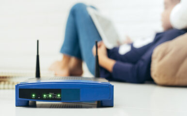 A close-up of a blue wireless router on a white surface, with its indicator lights glowing. In the background, a child is blurred while using a tablet. "Understanding how often you should replace your router is essential for maintaining optimal internet connectivity and performance."