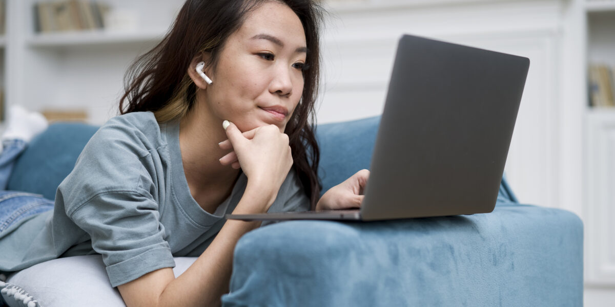 A young woman wearing wireless earbuds lies on a blue couch, using a laptop for online learning. She appears focused, possibly searching for solutions on how to fix slow WiFi while working from home.