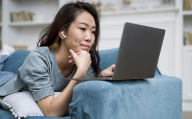 A young woman wearing wireless earbuds lies on a blue couch, using a laptop for online learning. She appears focused, possibly searching for solutions on how to fix slow WiFi while working from home.