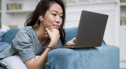 A young woman wearing wireless earbuds lies on a blue couch, using a laptop for online learning. She appears focused, possibly searching for solutions on how to fix slow WiFi while working from home.